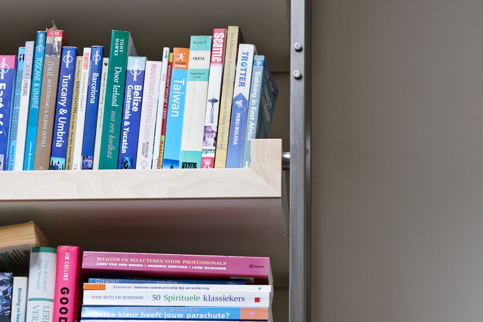 Detail van een houten boekenplank met reisgidsen, naast een stalen bevestigingsbout in de zijwand.
