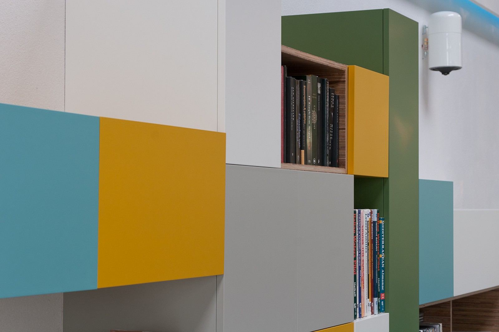 Detail van een kleurrijk kastenblok in turquoise, oker en donkergroen, met een boekennis tussen de vakken.