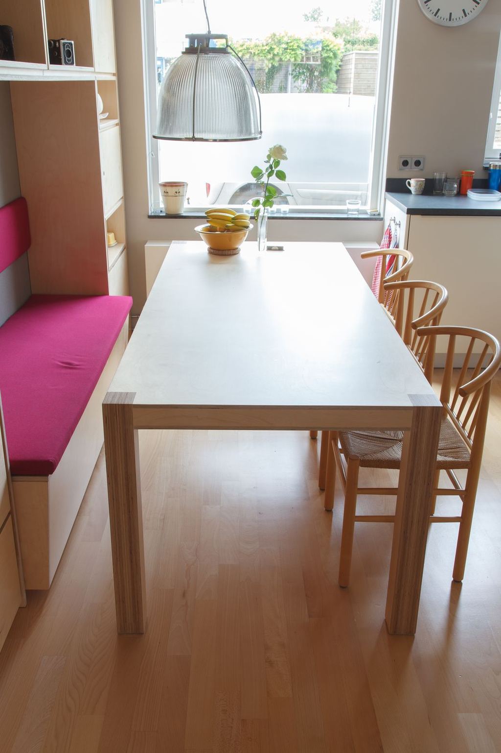 Eettafel met vlechtstoelen naast een roze zitbank en een houten kastwand.