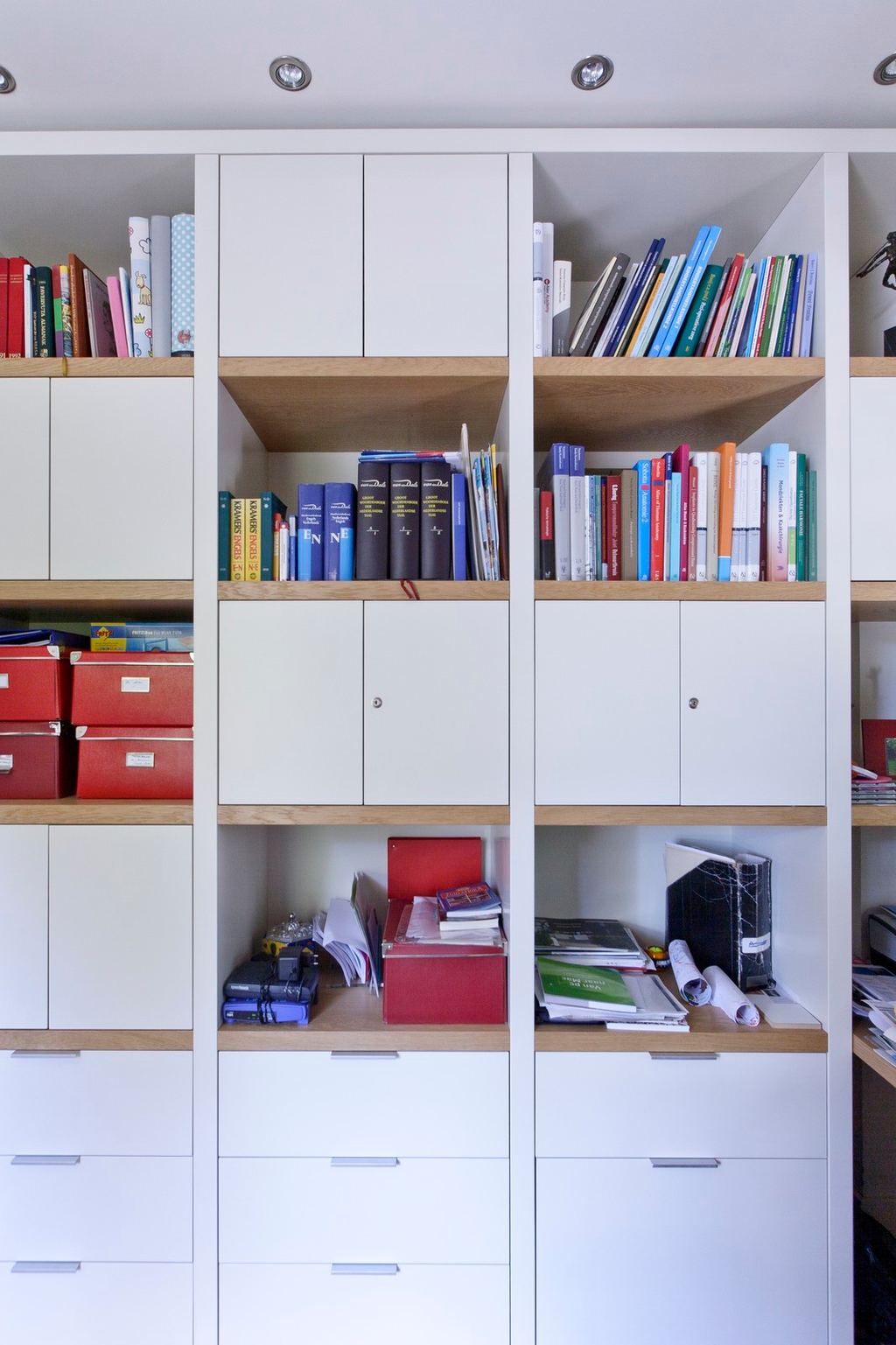 Witte wandkast met eiken planken vol boeken en rode dozen.