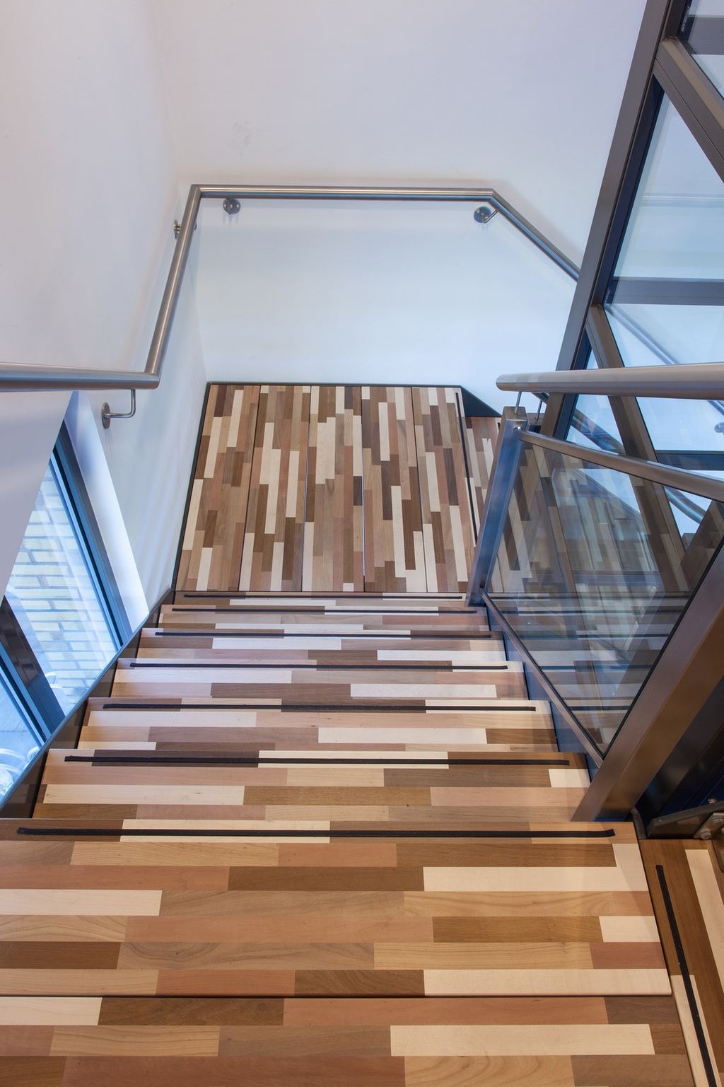 Glazen balustrade langs de trap met houten treden in verschillende kleuren.