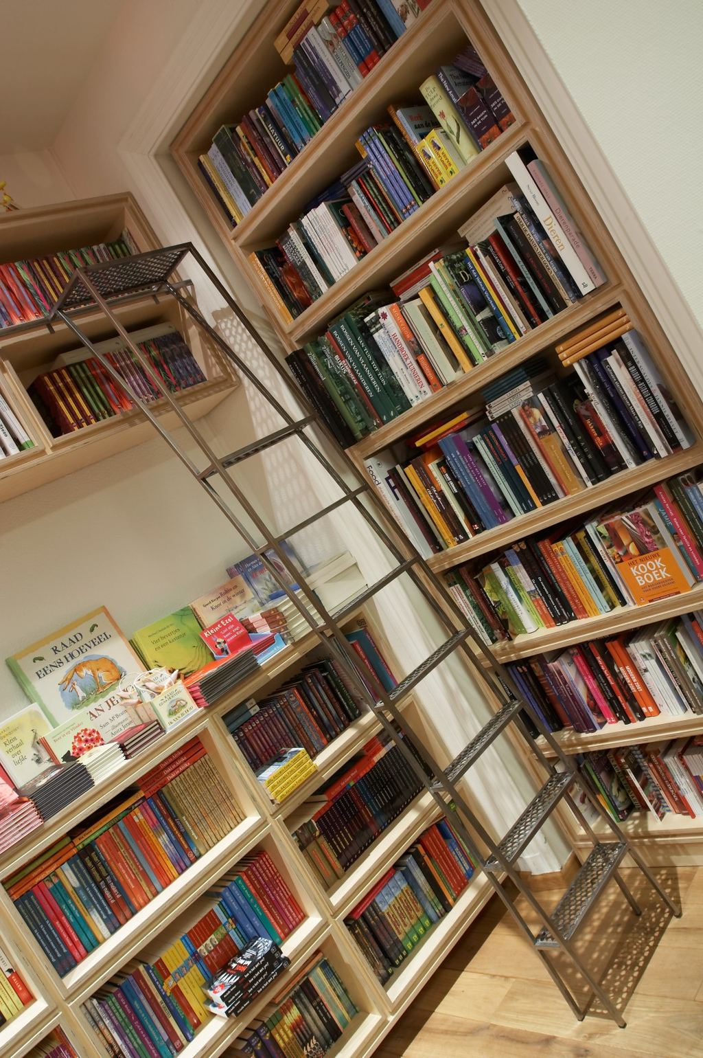 Hoekboekenkast van berken multiplex die de aflopende zoldervorm volgt, met een ladder.