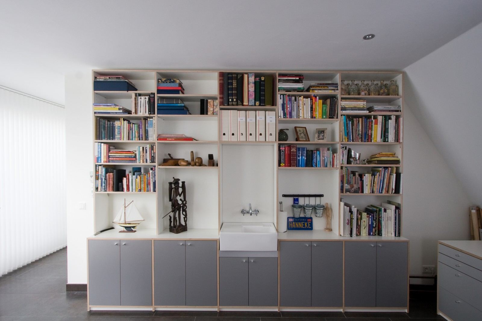 Wandvullende boekenkast met een ingebouwde spoelbak boven grijze kastdeuren.