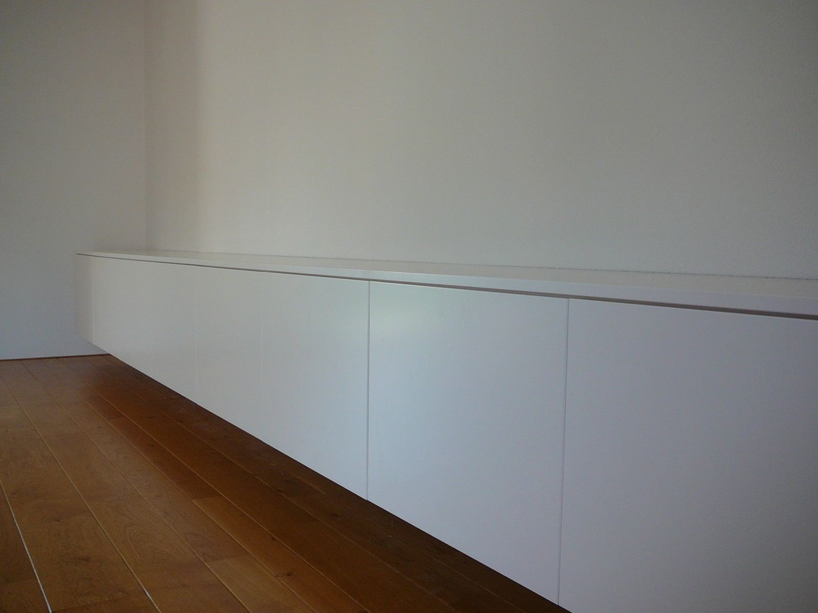 Extreem lange zwevende kast met witte, greeploze deuren tegen een lege wand.