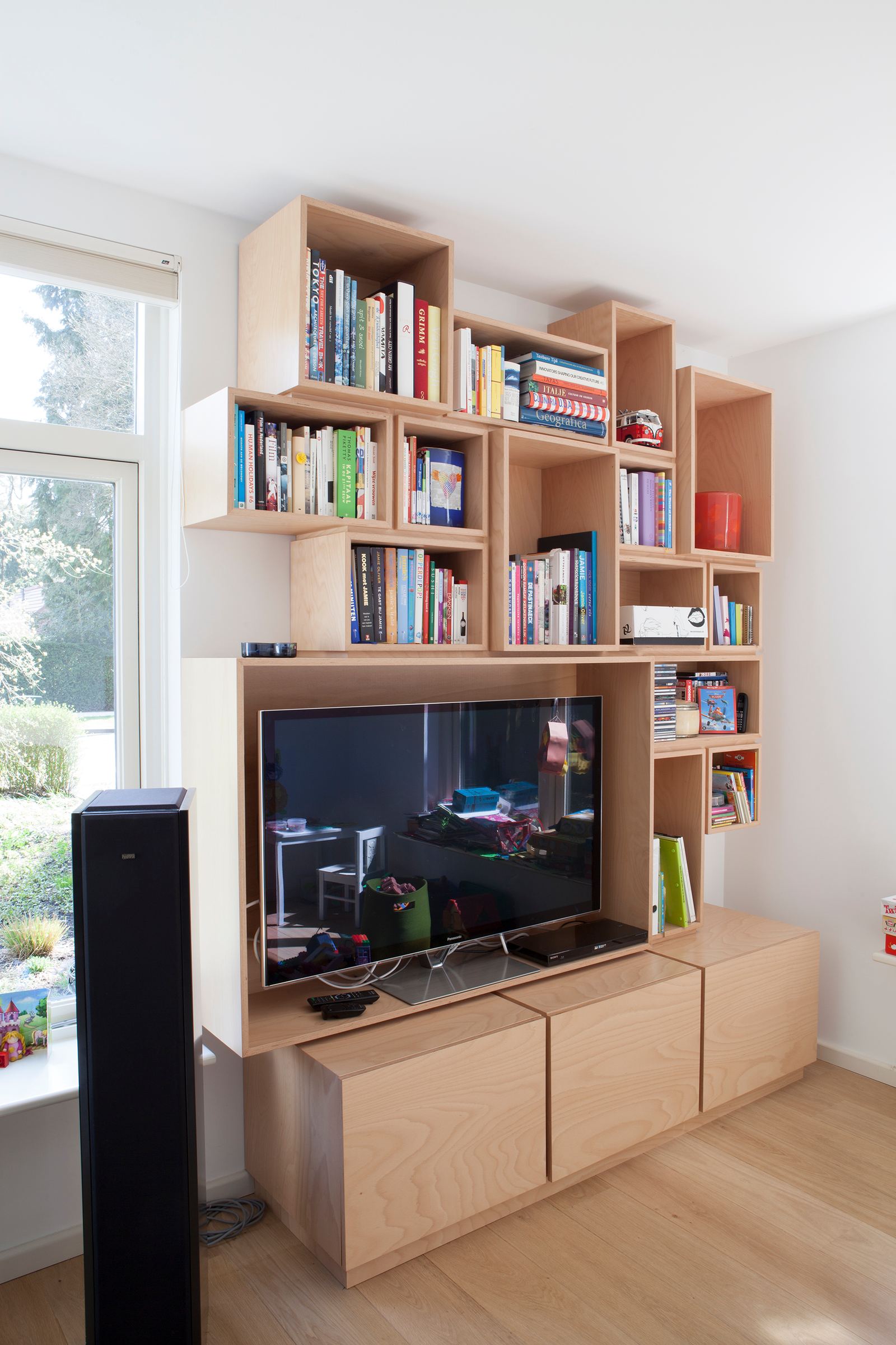 Puzzelvormige wandkast met losse vakken vol boeken, boven een tv-meubel met laden.